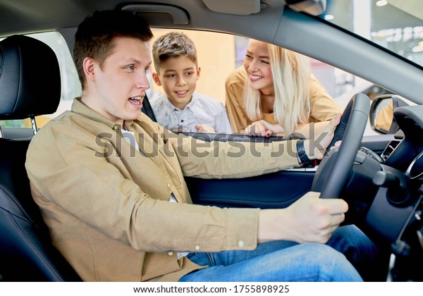 young caucasian man tries to drive a new car,\
his wife and child boy waiting for his reaction and feelings about\
car. in dealership