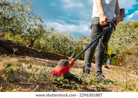 a young caucasian man using a cordless electric string trimmer to mow the grown grass of a farmland