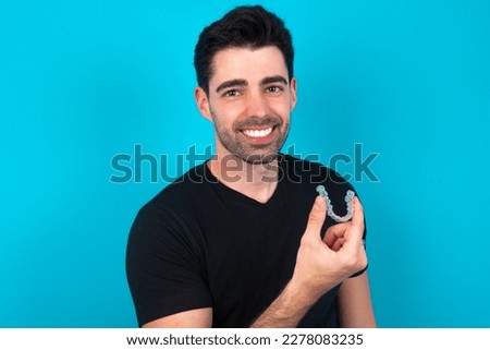 Young caucasian man wearing black T-shirt over blue background holding an invisible aligner ready to use it. Dental healthcare and confidence concept.