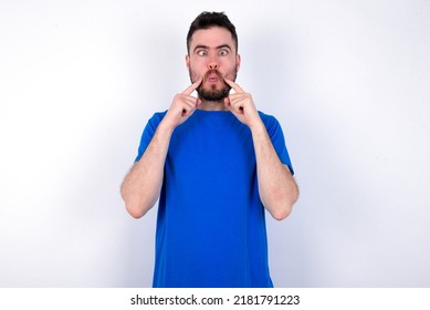 Young caucasian man wearing blue T-shirt over white background crosses eyes and makes fish lips funny grimace