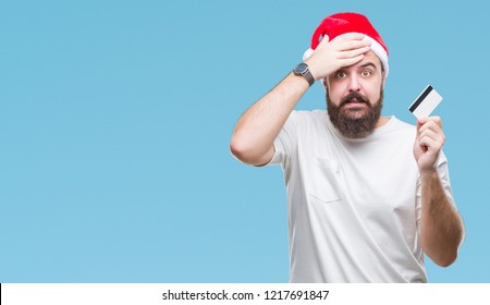 Young caucasian man wearing christmas hat holding credit card over isolated background stressed with hand on head, shocked with shame and surprise face, angry and frustrated. Fear and upset Stock fotografie