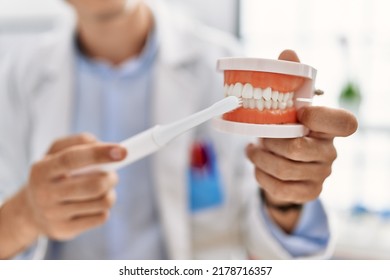 Young Caucasian Man Wearing Dentist Uniform Cleaning Denture Using Toothbrush At Clinic