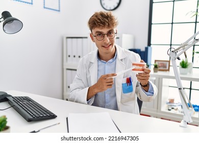 Young Caucasian Man Wearing Dentist Uniform Cleaning Denture Using Toothbrush At Clinic