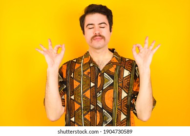 Young caucasian man wearing generic pattern printed shirt against yellow wall doing yoga, keeping eyes closed, holding fingers in mudra gesture. Meditation, religion and spiritual practices. - Powered by Shutterstock