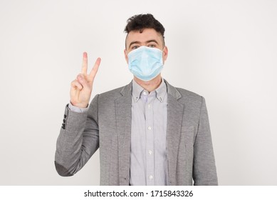 Young caucasian man wearing medical mask standing against gray wall showing and pointing up with fingers number two while smiling confident and happy.