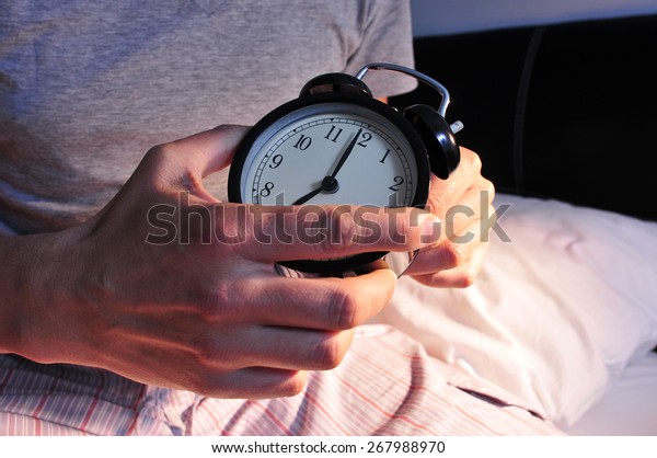 a young caucasian man wearing\
pajamas in bed setting the alarm clock at 7 before lie\
down