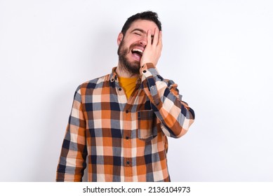 Young Caucasian Man Wearing Plaid Shirt Over White Background Makes Face Palm And Smiles Broadly, Giggles Positively Hears Funny Joke Poses