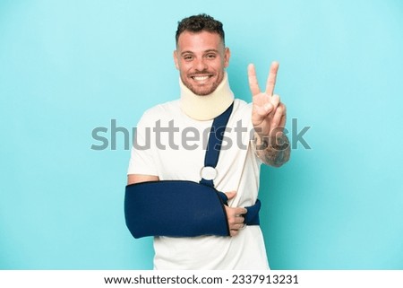 Young caucasian man wearing a sling  and neck lace isolated on blue background smiling and showing victory sign