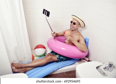 a young caucasian man wearing sunglasses, a straw hat and a pink swim ring in a blue air mattress placed in the shower of a bathroom takes a selfie with his smartphone while is drinking a red cocktail
