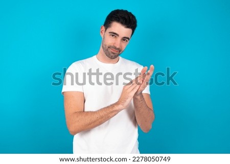 Young caucasian man wearing white T-shirt over blue background clapping and applauding happy and joyful, smiling proud hands together.