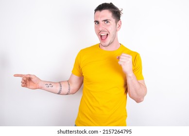 Young caucasian man wearing yellow t-shirt over white background points at empty space holding fist up, winner gesture. - Shutterstock ID 2157264795