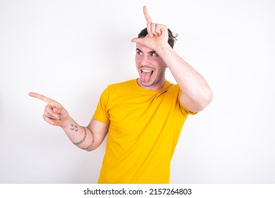 Young caucasian man wearing yellow t-shirt over white background showing loser sign and pointing at empty space - Shutterstock ID 2157264803