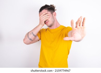 Young caucasian man wearing yellow t-shirt over white background covers eyes with palm and doing stop gesture, tries to hide. Don't look at me, I don't want to see, feels ashamed or scared.