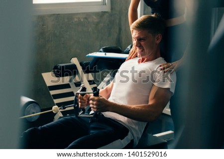 Young Caucasian man in white shirt at a gym, training hard and pulling weights in seated cable row machine. Lifting heavy weights and fatigue. Sport fitness and muscles concept