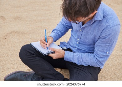 Young Caucasian Man Writing A Letter Or Poetry In His Notebook, Sitting On The Sand On The Beach, In Formal Attire.