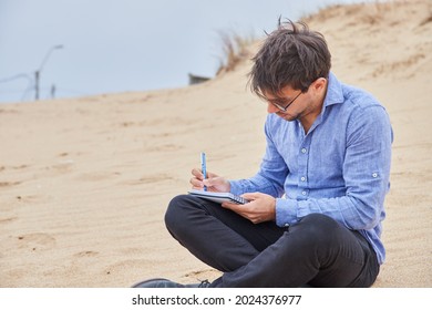 Young Caucasian Man Writing A Letter Or Poetry In His Notebook, Sitting On The Sand On The Beach, In Formal Attire.