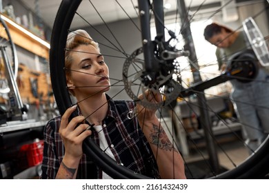 Young Caucasian Mechanic Girl Installing Bike Cassette On Wheel While Her Blurred Black Female Colleague Fixing Electric Scooter On Background In Modern Workshop. Bike Service, Repair And Upgrade