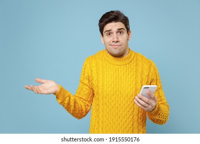 Young Caucasian Mistaken Confused Student Man 20s In Casual Knitted Yellow Sweater Hold In Hand Using Mobile Cell Phone Spreading Hand Oops Emotion Isolated On Blue Color Background Studio Portrait