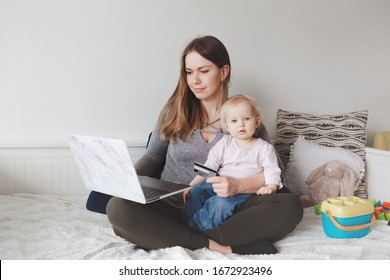 Young Caucasian Mother With Baby Shopping Online From Home. Stay At Home Single Mom With Kid Toddler Daughter Buying Ordering Products Food On Internet And Paying With Credit Card. 