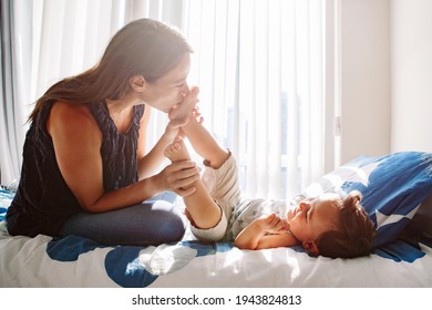 Young Caucasian Mother Kissing Boy Toddler Son Feet Heels Legs. Mom Playing With Child Baby On Bed In Bedroom At Home. Happy Authentic Family Childhood Lifestyle. Tender Touching Sweet Moments.