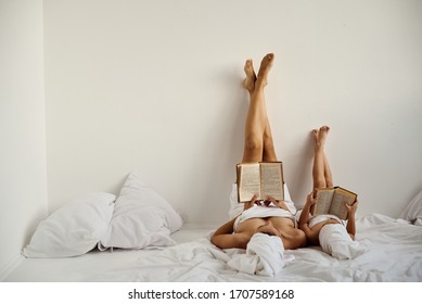 A Young Caucasian Mother And Little Daughter With Wrapped Hair In White Bath Towels Read Books Lying On The Bed And Lifting Their Feet Up Against The White Wall. 