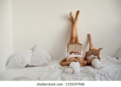 A Young Caucasian Mother And Little Daughter With Wrapped Hair In White Bath Towels Read Books Lying On The Bed And Lifting Their Feet Up Against The White Wall. 