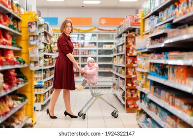 A young caucasian mother rolls a grocery cart with a baby sitting in it through the supermarket. Indoor. The concept of family shopping.