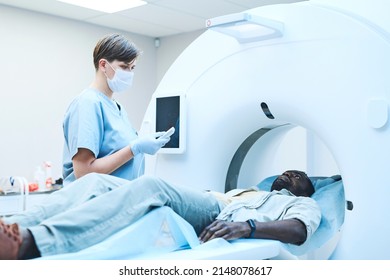 Young Caucasian MRI Tech In Mask Pushing Button On Control Panel While Preparing Patient For Scanning