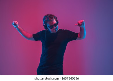 Young caucasian musician in headphones singing on gradient pink-purple background in neon light. Concept of music, hobby, festival. Joyful party host, DJ, stand upper. Colorful portrait of artist.