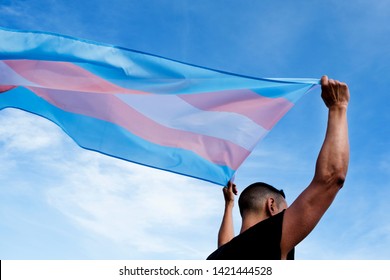 a young caucasian person, seen from behind, holding a transgender pride flag over his or her head against the blue sky