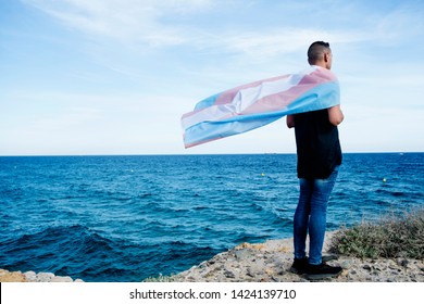 a young caucasian person, seen from behind, draping a transgender pride flag over his or her shoulders, facing the ocean