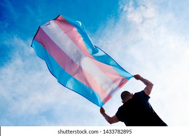 eine junge, kaukasische Person, die von hinten gesehen wird und eine transgender-Stolz-Flagge über ihrem Kopf gegen den blauen Himmel trägt