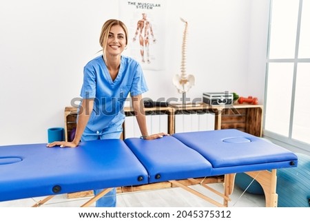 Young caucasian physio therapist smiling happy leaning on table at the clinic