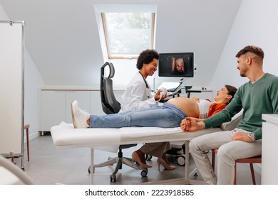 A young Caucasian pregnant woman is on the table at the doctor's office having an ultrasound checkup by an African American doctor while her husband is there for emotional support. - Shutterstock ID 2223918585