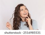 Young caucasian pretty brunette woman with a curly hair and wearing a blue t-shirt looks up, dreaming, smiling and holding a strand of hair in her hand. Isolated on a light background. 