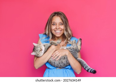A young caucasian pretty cute blonde woman in a blue dress holds a cat in her hands like a baby isolated on a bright color pink background. The girl babying with a kitten. Fiendship of pet and owner