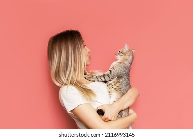 A young caucasian pretty cute blonde woman holds a tabby cat in her hands admiring it isolated on a bright color pink background. The girl babying with a kitten. Fiendship of pet and owner. Banner
