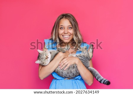 A young caucasian pretty cute blonde woman in a blue dress holds a cat in her hands like a baby isolated on a bright color pink background. The girl babying with a kitten. Fiendship of pet and owner
