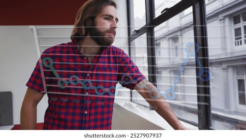 A young Caucasian professional wearing red plaid shirt, gazing out a window. Sporting long brown hair and a beard, standing in a modern office with a white background - Powered by Shutterstock