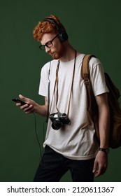 Young Caucasian Red Hair Guy With Photo Camera And Backpack Listen Music In Earphones On Smartphone. Concept Of Human Of Generation Z. Male Traveler Look At Camera On Dark Green Background In Studio