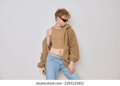 Young caucasian red head queer gender person with a bold trendy look wearing sunglasses, beige teddy coat, blue low-waist jeans, brown crop top and silver belt posing isolated over grey background. - Powered by Shutterstock