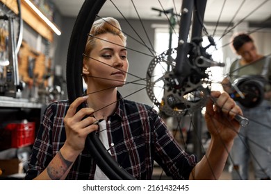 Young caucasian repairman girl installing bike cassette on wheel while her blurred black female colleague fixing electric scooter on background in modern workshop. Bike service, repair and upgrade - Shutterstock ID 2161432445