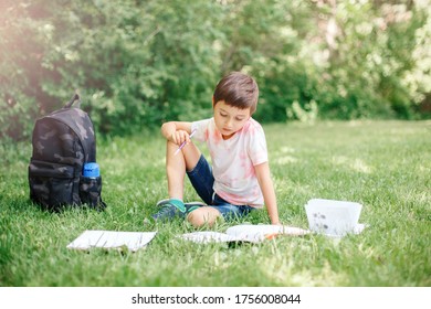 Young Caucasian School Boy Sitting In Park Outdoor Doing School Homework. Child Kid Writing In Notebook With Pencil Outside. Self Education Learning Studying. Early Development For Children.