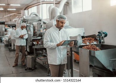 Young Caucasian Serious Supervisor Evaluating Quality Of Food In Food Plant While Holding Tablet. Man Is Dressed In White Uniform And Having Hair Net.