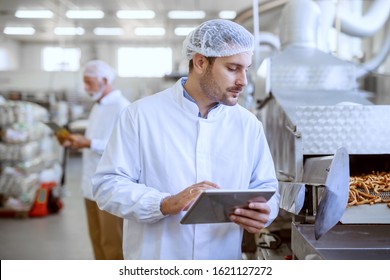 Young Caucasian Serious Supervisor Evaluating Quality Of Food In Food Plant While Holding Tablet. Man Is Dressed In White Uniform And Having Hair Net.