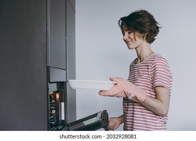 Young Caucasian Side View Housewife Woman 20s In Casual Clothes Striped T-shirt Puts Casserole In Baking Dish In Oven Cooking Food In Light Kitchen At Home Alone. Healthy Diet Bakery Lifestyle Concept