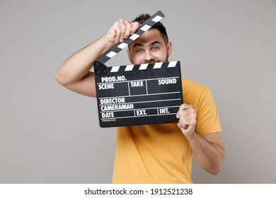 Young caucasian smiling bearded attractive cameramen producer happy man 20s in casual yellow basic t-shirt covering face with clapperboard isolated on grey background studio portrait. Cinema concept