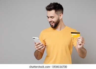 Young Caucasian Smiling Bearded Attractive Rich Student Man 20s Wear Casual Yellow Basic T-shirt Hold Mobile Cell Phone Credit Bank Card Shopping Online Isolated On Grey Background Studio Portrait