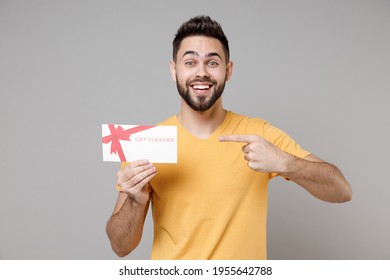 Young Caucasian Smiling Bearded Attractive Handsome Happy Man 20s Wearing Casual Yellow Basic T-shirt Point Index Finger On Gift Voucher Flyer Mock Up Isolated On Grey Color Background Studio Portrait