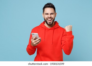 Young Caucasian Smiling Bearded Man 20s Wearing Casual Red Orange Hoodie Hold Mobile Cell Phone Text Do Winner Gesture Clench Fist Isolated On Blue Background Studio Portrait Modern Technology Concept